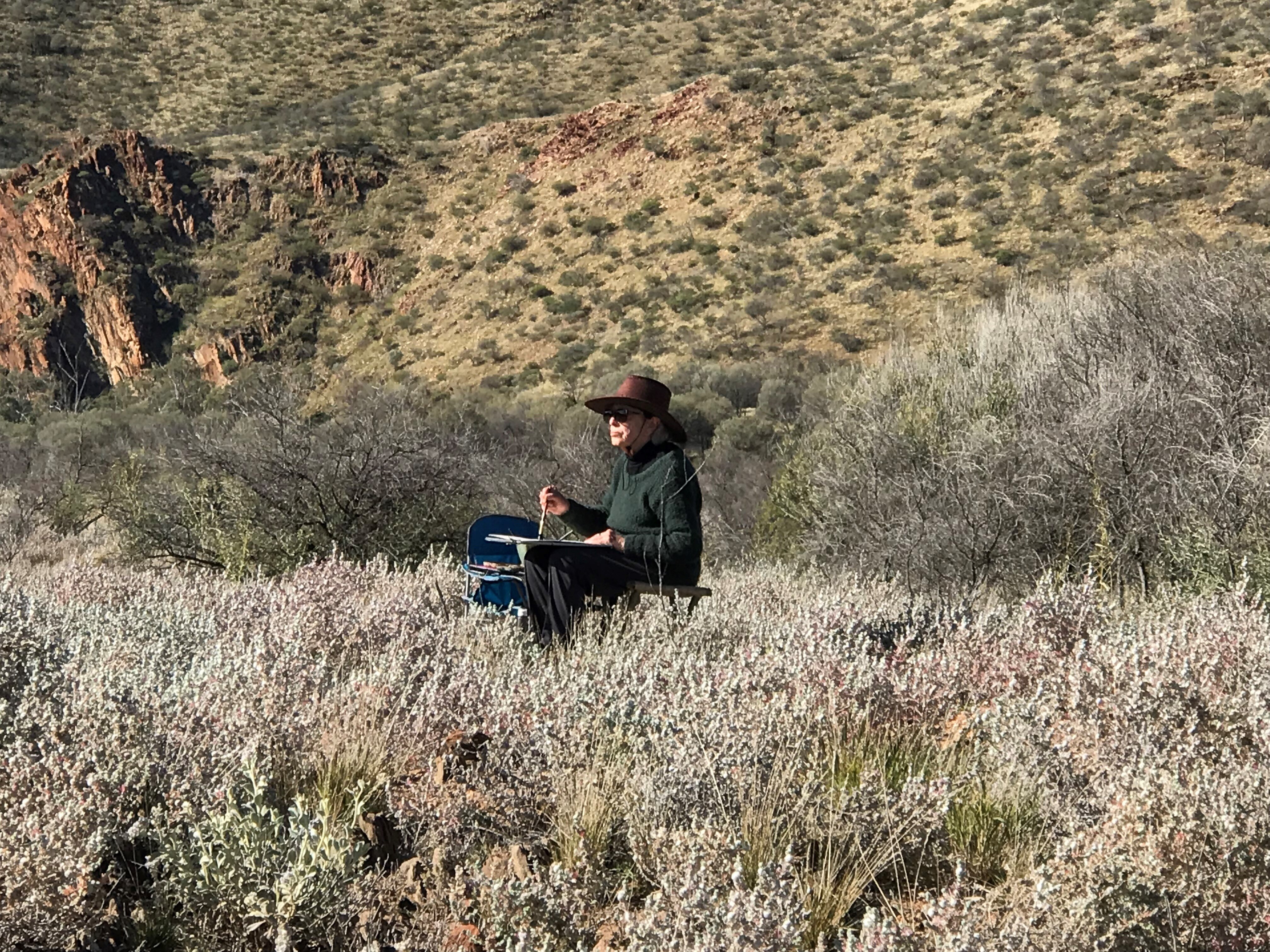 A woman with a wide brim hat sits in a camp chair holding a paint brush surrounded by bush. 