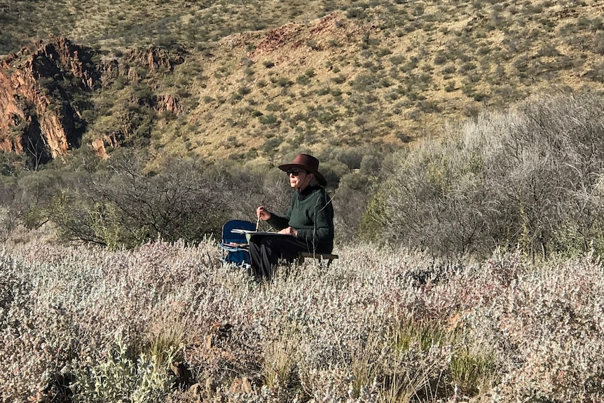 A woman with a wide brim hat sits in a camp chair holding a paint brush surrounded by bush. 