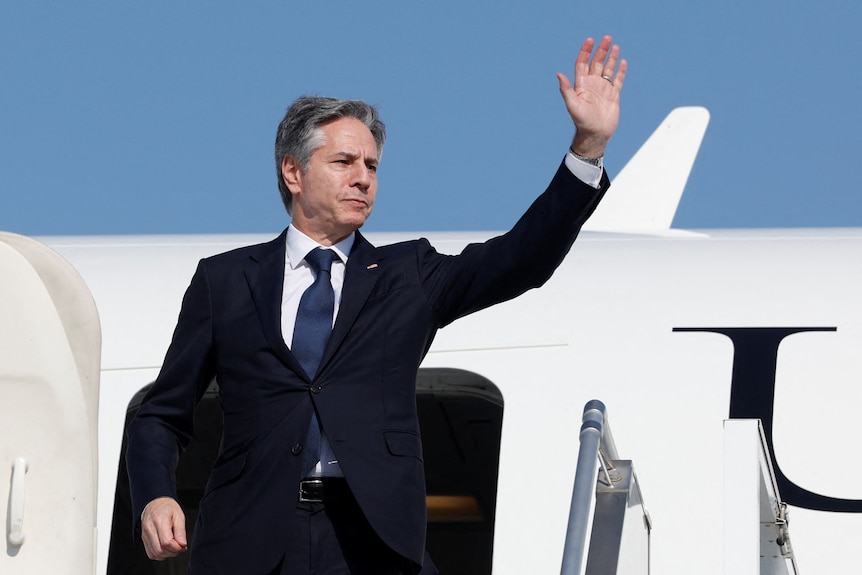 A middle-aged man in a suit waves against a blue-sky backdrop as he boards a white plane.
