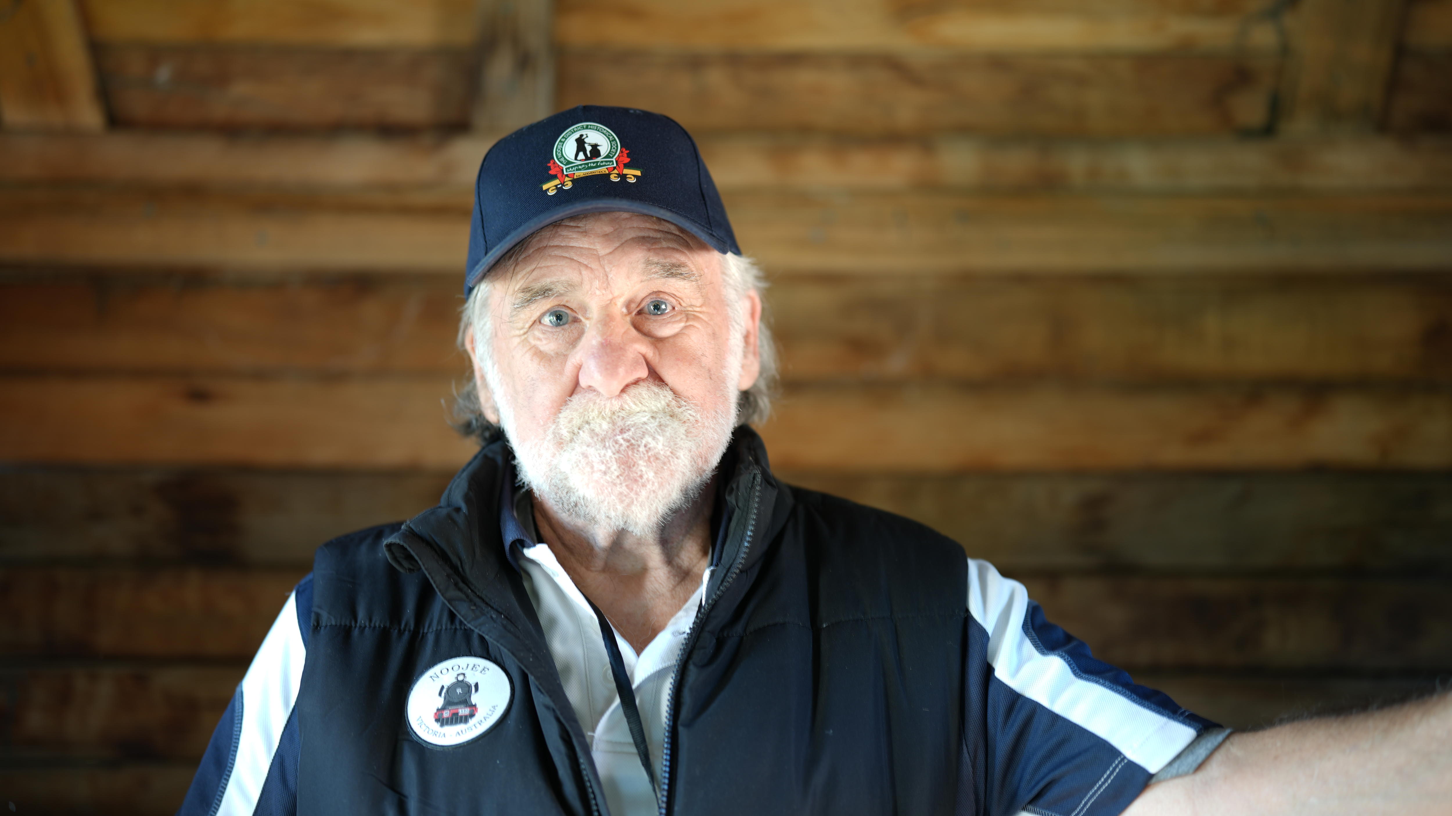 Image of a man with a white bear and a navy baseball cap, with a timber background. 