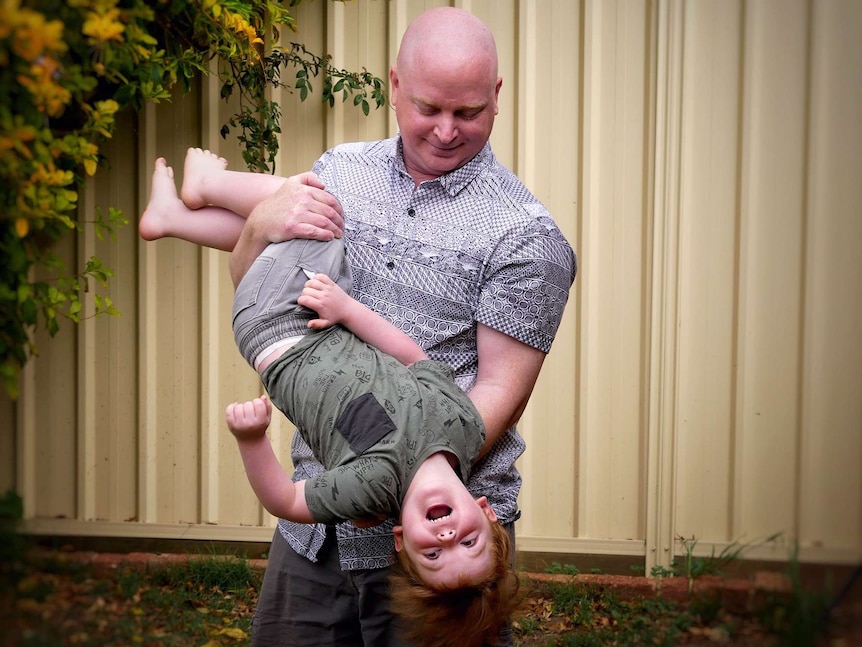 A man stands smiling in a backyard holding a laughing young boy upside down.