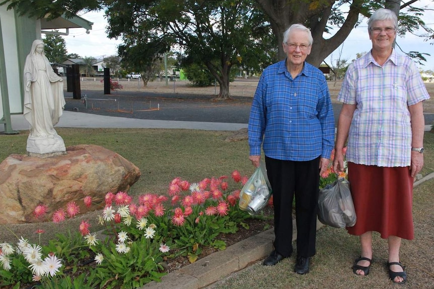 Two elderly women stand by a garden at a church holding bags of rubbish