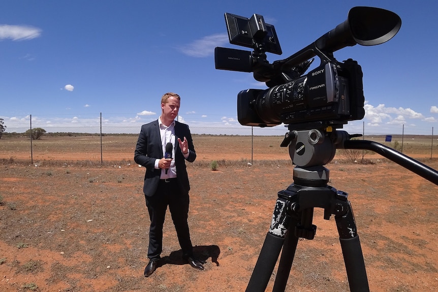 A man in a suit standing in front of a camera holding a microphone in a dirt field