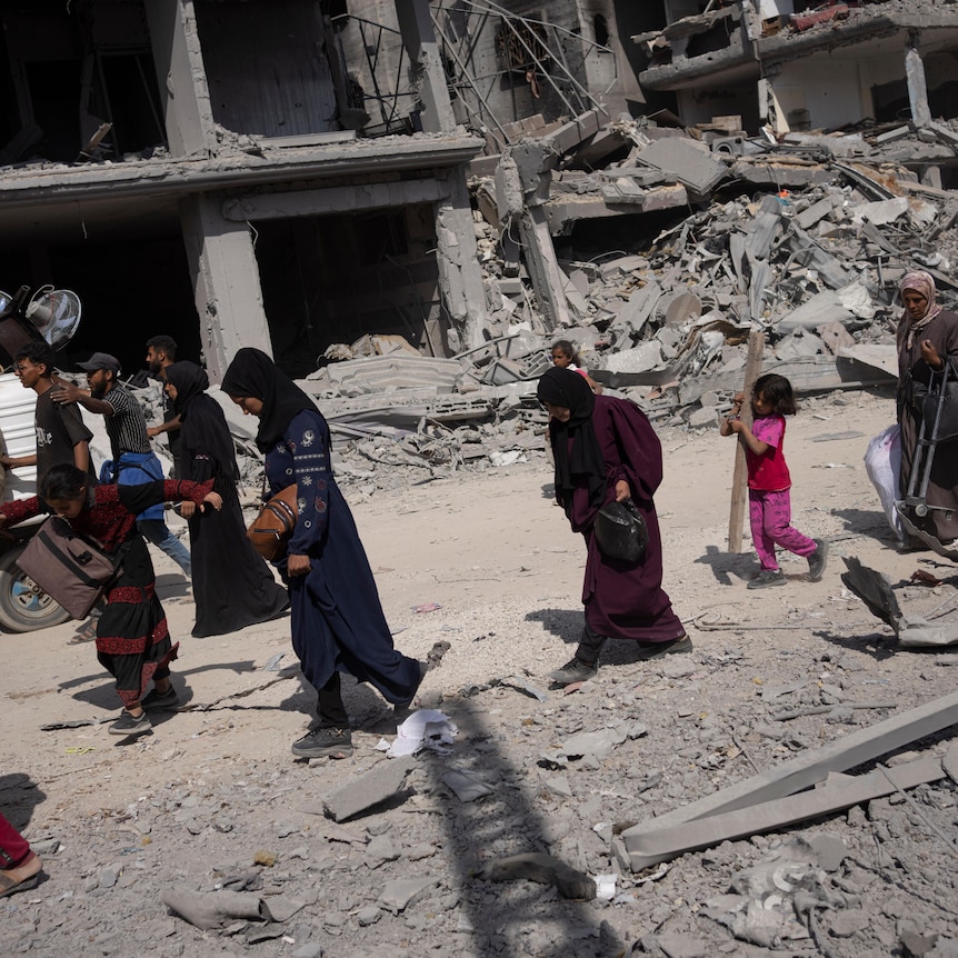A group of people including young children and women walk down a street amid rubble and bombed buildings