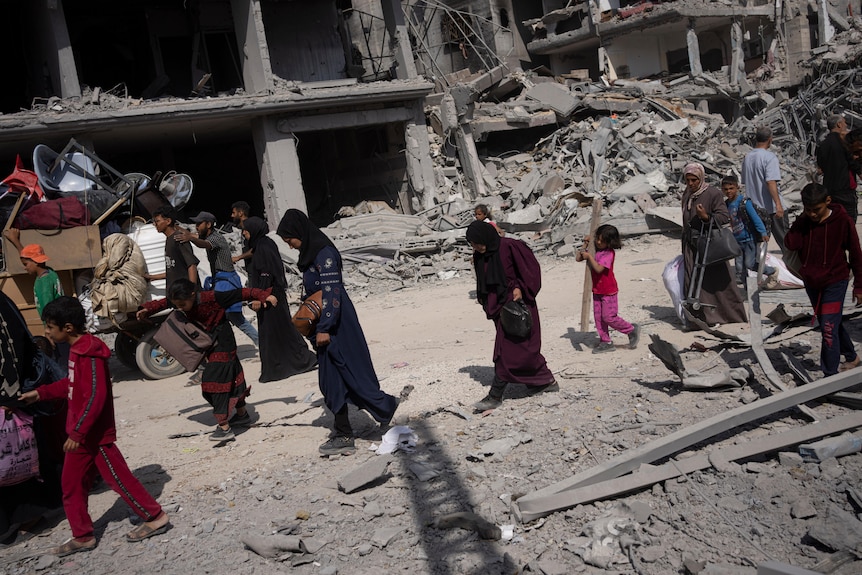 A group of people including young children and women walk down a street amid rubble and bombed buildings