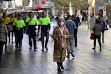 A woman wears a mask in an outside mall while other people walk behind her without one