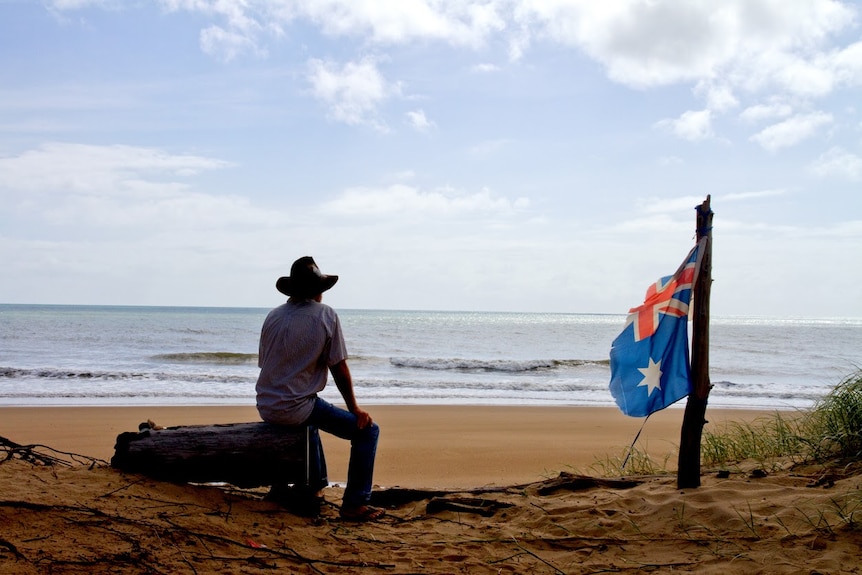 A man in a hat sits looking over a beach with deteriorated Australian flag flying next to him