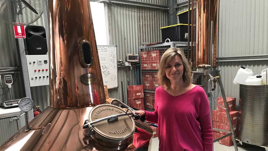 A woman smiles as she leans on a large copper still inside a large shed.