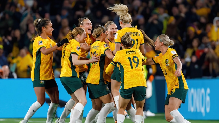 Female soccer players in gold jerseys embraces in celebration after scoring a goal