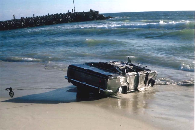 A washed up car is dragged out of the water at a Perth beach, with a rocky groyne in the background.