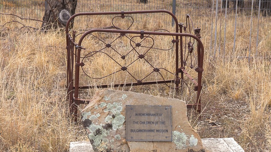 A plaque on a rock in front of a rusted old fashioned cot set in dry grassland