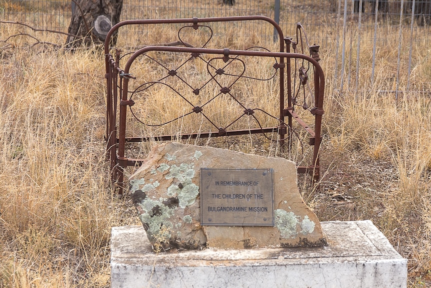 A plaque on a rock in front of a rusted old fashioned cot set in dry grassland