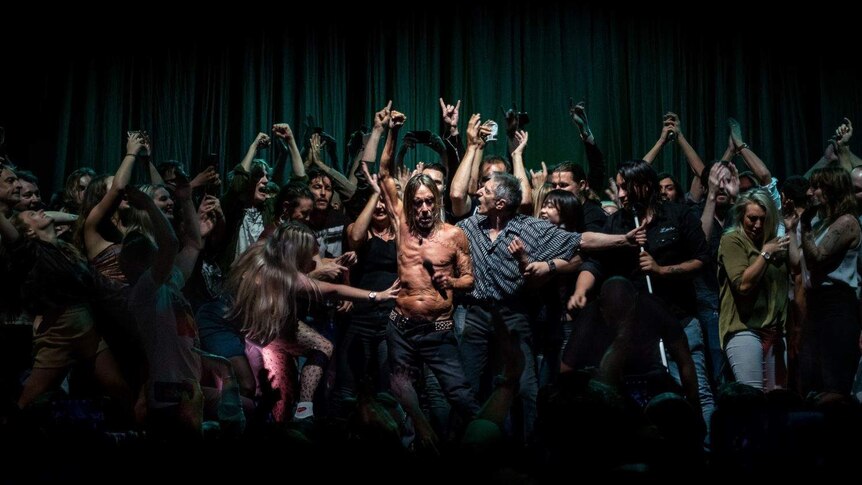 Shirtless Iggy Pop holding microphone surrounded by dancing fans