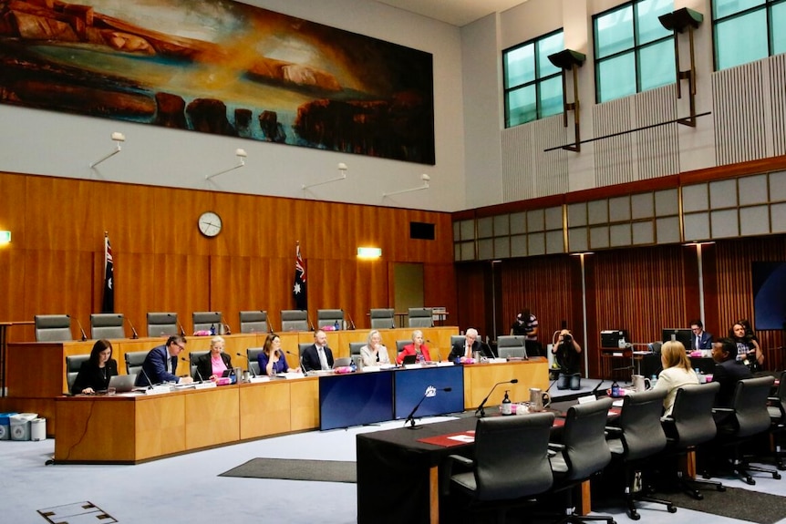 A woman sitting on a bench in front of a long table of people at a senate hearing inside a building in Canberra