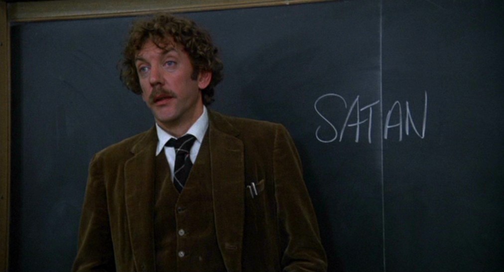 A man in a tweed suit stands in front of a blackboard with the word 'SATAN' written on it.