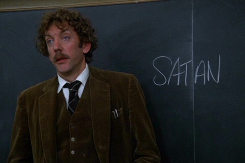 A man in a tweed suit stands in front of a blackboard with the word 'SATAN' written on it.