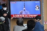A small crowd of people watch a TV set with a missile launch as a police officer stands next to the monitor