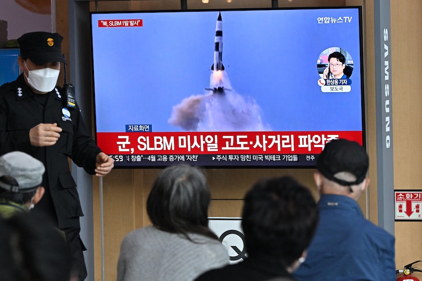 A small crowd of people watch a TV set with a missile launch as a police officer stands next to the monitor