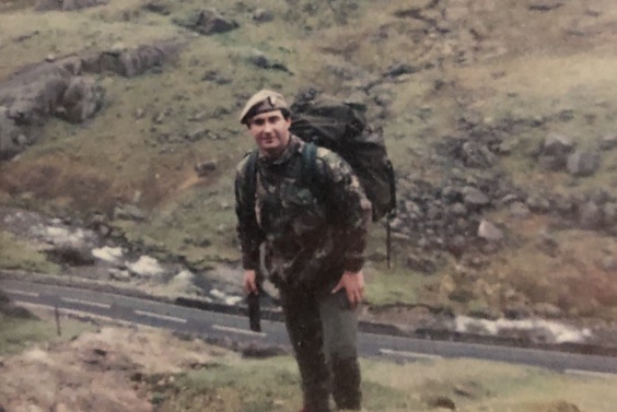 A soldier stands on a grassy hill and smiles at the camera