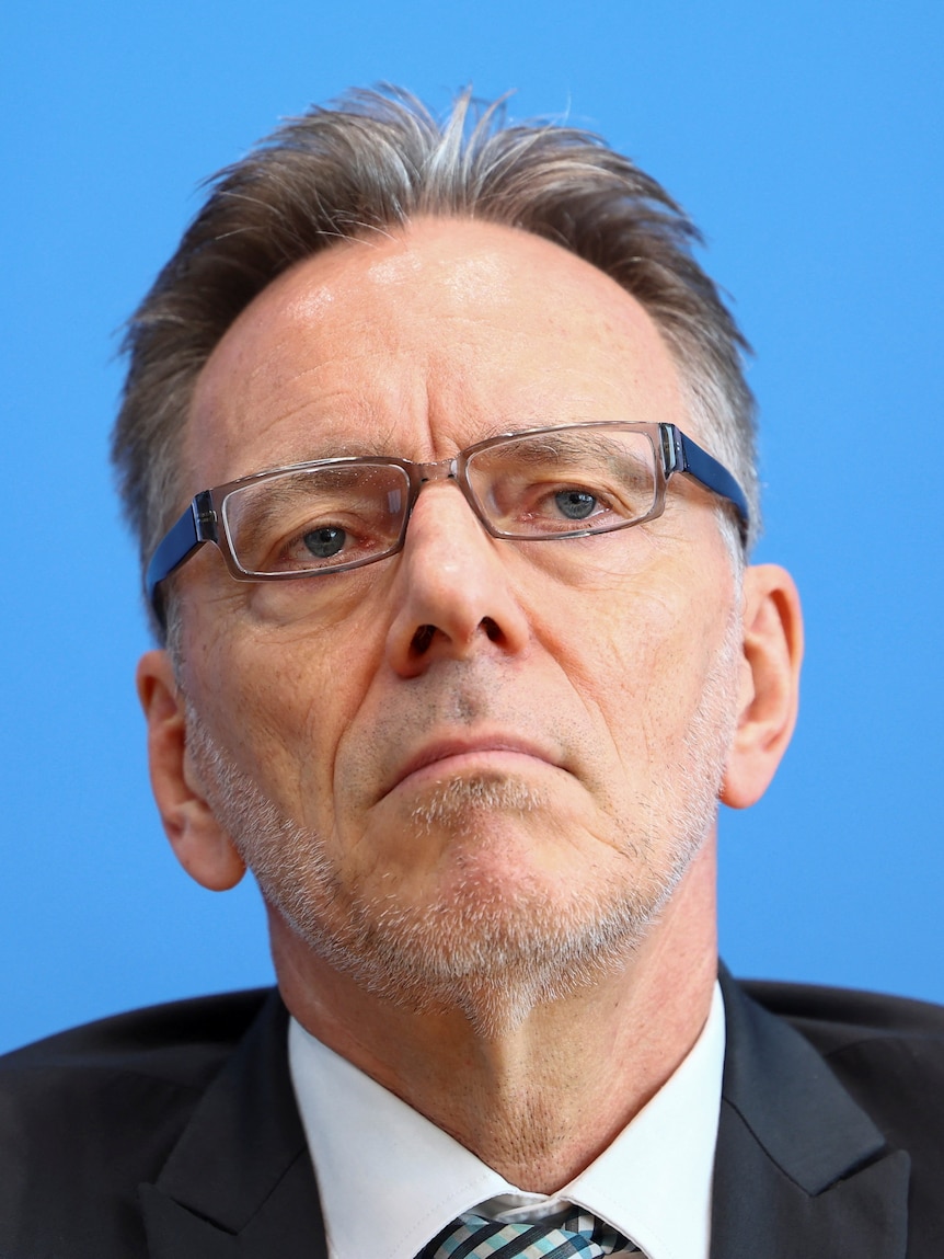 Holger Muench, an older man with glasses and a suit, sits in front of a blue background. 