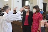 A priest raises his hands over two women in a church, all three are wearing face masks over their noses and mouths. 