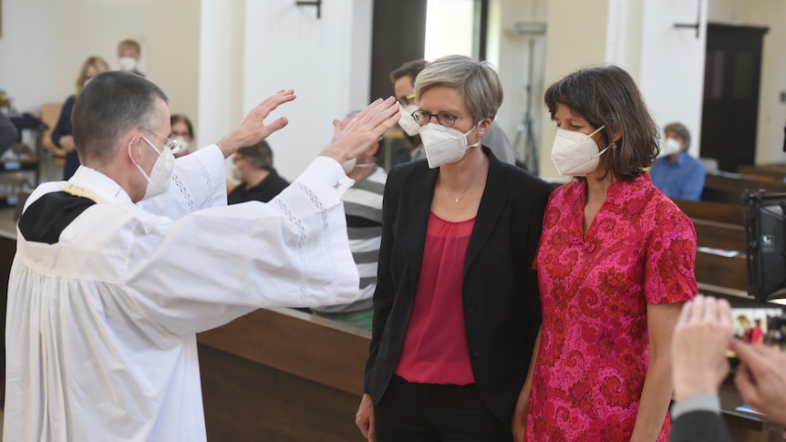 A priest raises his hands over two women in a church, all three are wearing face masks over their noses and mouths. 