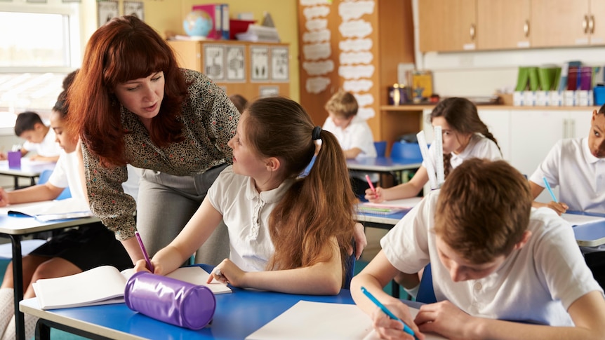 A female teacher with red hair leans down to the desk of a primary-school student to help her with a question in class.