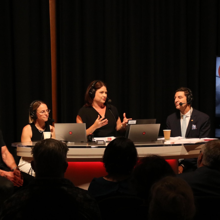 Four panellists with microphones sit behind a desk in a radio studio with a live audience watching on