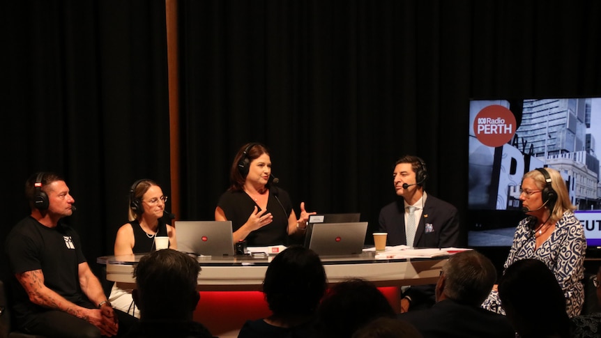 Four panellists with microphones sit behind a desk in a radio studio with a live audience watching on