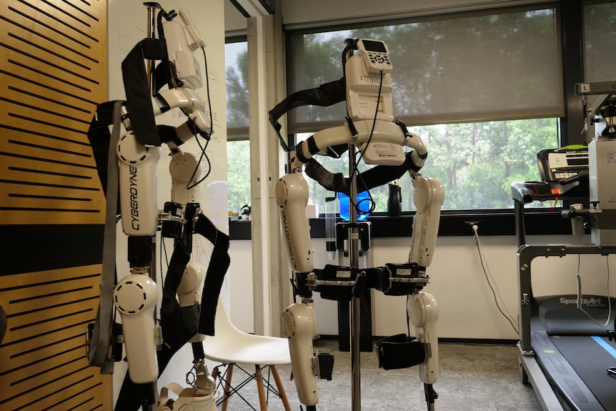 Two white robotic exoskeletons stand alongside each other, supported by metal poles.