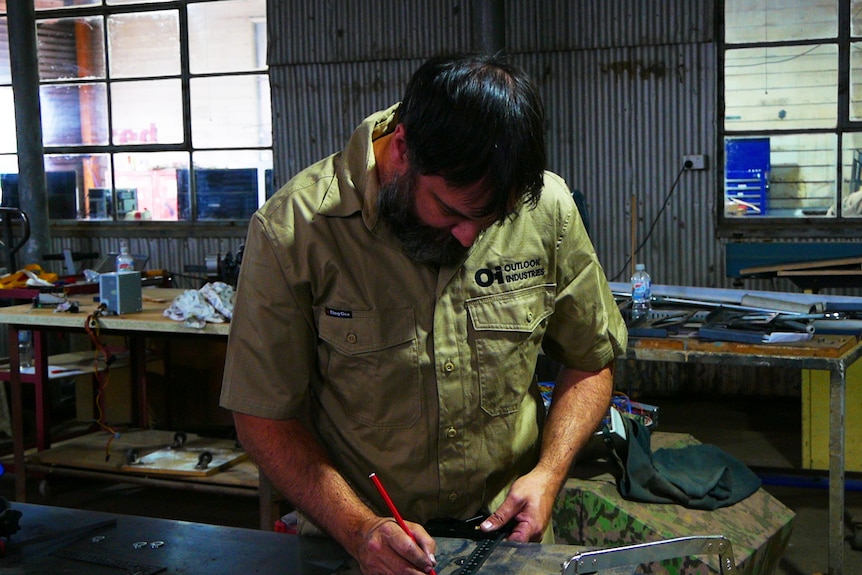 A man in a khaki shirt in a workshop reviewing his work with a pencil in hand