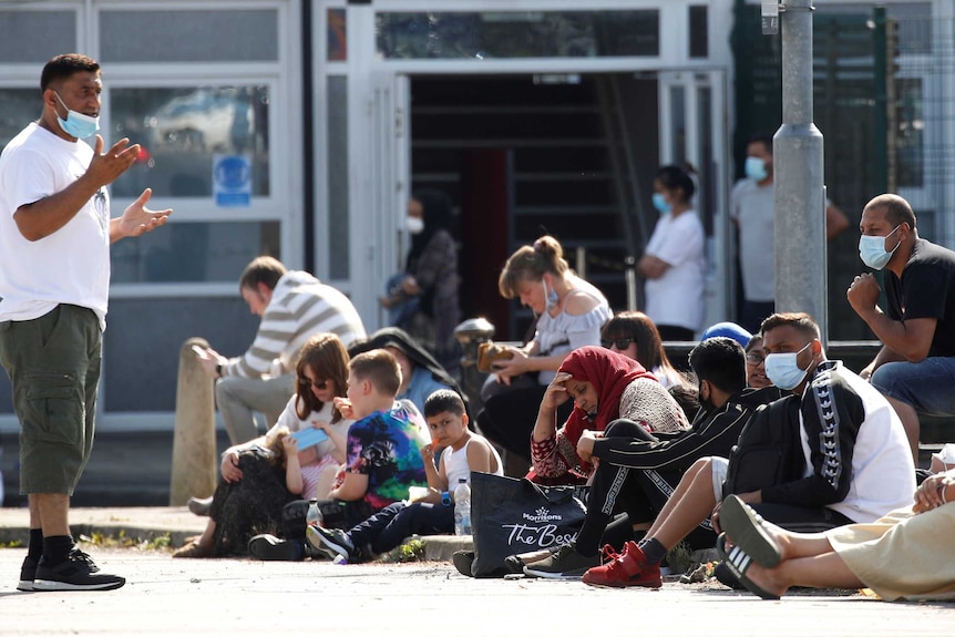 One man wearing a mask gesticulates to others, who are seated in the gutter as they wait for coronavirus tests in Bury, UK.