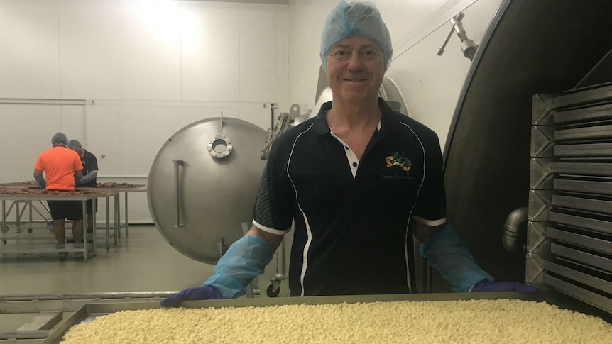Michael Buckley smiles at the camera with a tray of freeze dried cheese in the factory and workers in the distance behind him.
