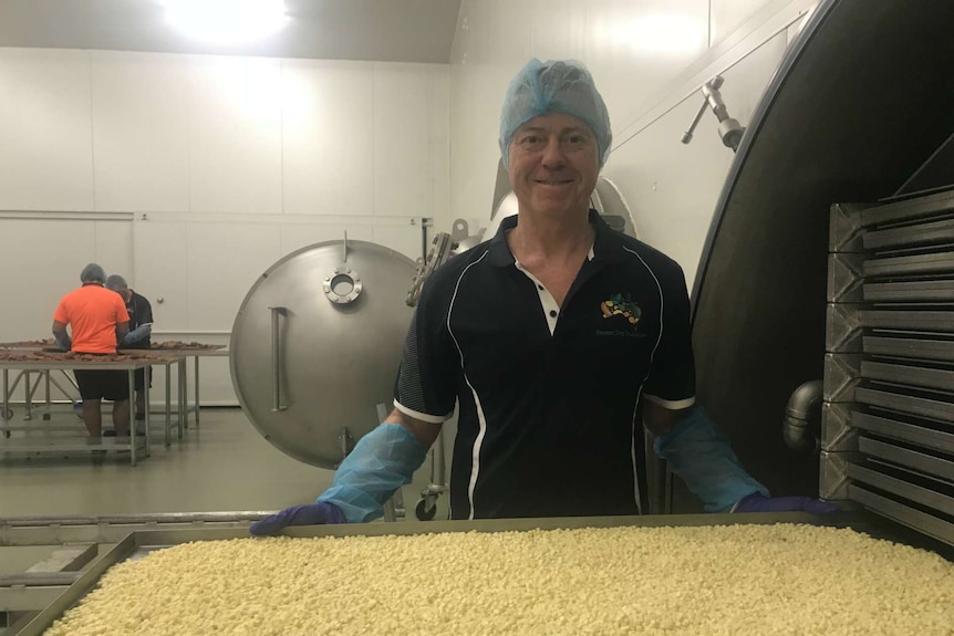 Michael Buckley smiles at the camera with a tray of freeze dried cheese in the factory and workers in the distance behind him.