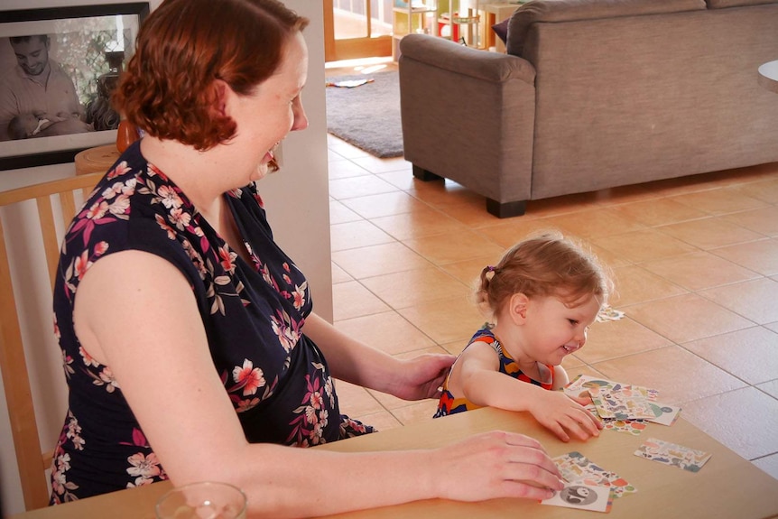 Pregnant woman with red hair wearing a floral dress playing a card game with young daughter at kitchen table.