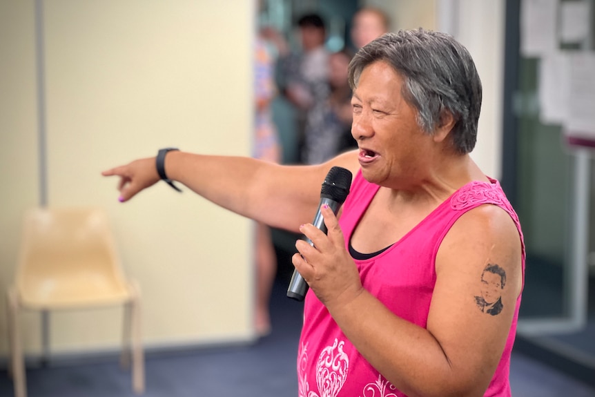 A woman in a pink tshirt with a tattoo of Elvis on her arm sings into a microphone.