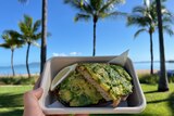 hand holds avocado toast with a tropical background