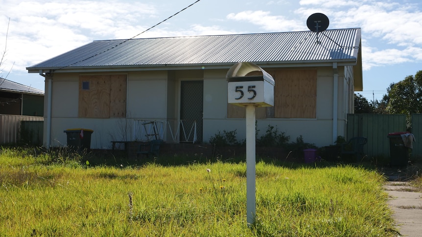 An abandoned house with boarded up windows and long grass.