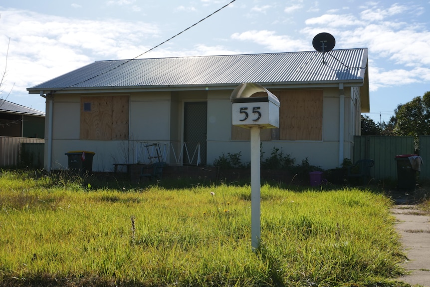 An abandoned house with boarded up windows and long grass.