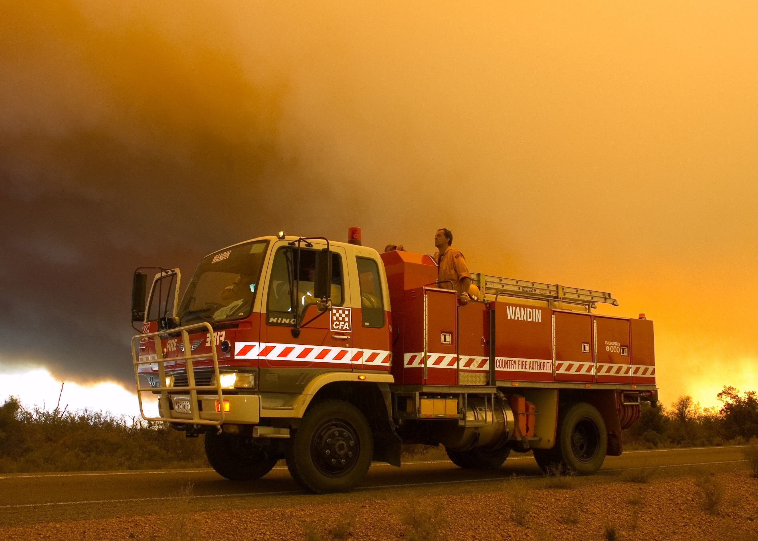 A man sits atop a CFA truck with a large orange smoke cloud behind it 