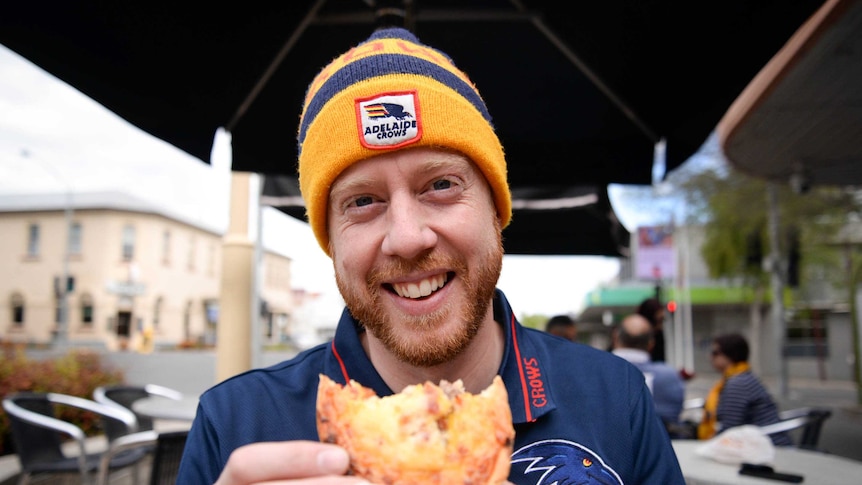 A man wearing an Adelaide Crows beanie smiles as he holds a pie while standing on the street.