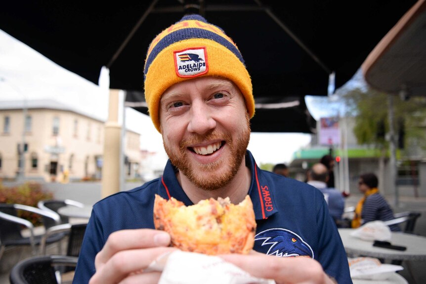 A man wearing an Adelaide Crows beanie smiles as he holds a pie while standing on the street.