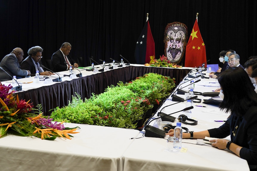 Three men in suits sit across a table from a diplomatic team of five people in a formal meeting with PNG and Chinese flags.