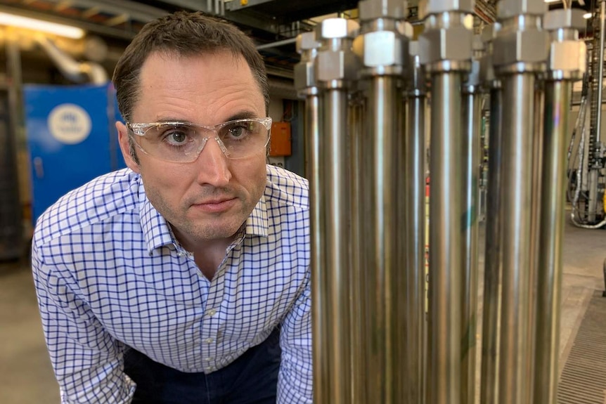 A man stands in a laboratory with clear glasses over his eyes and looks at metal pipes.