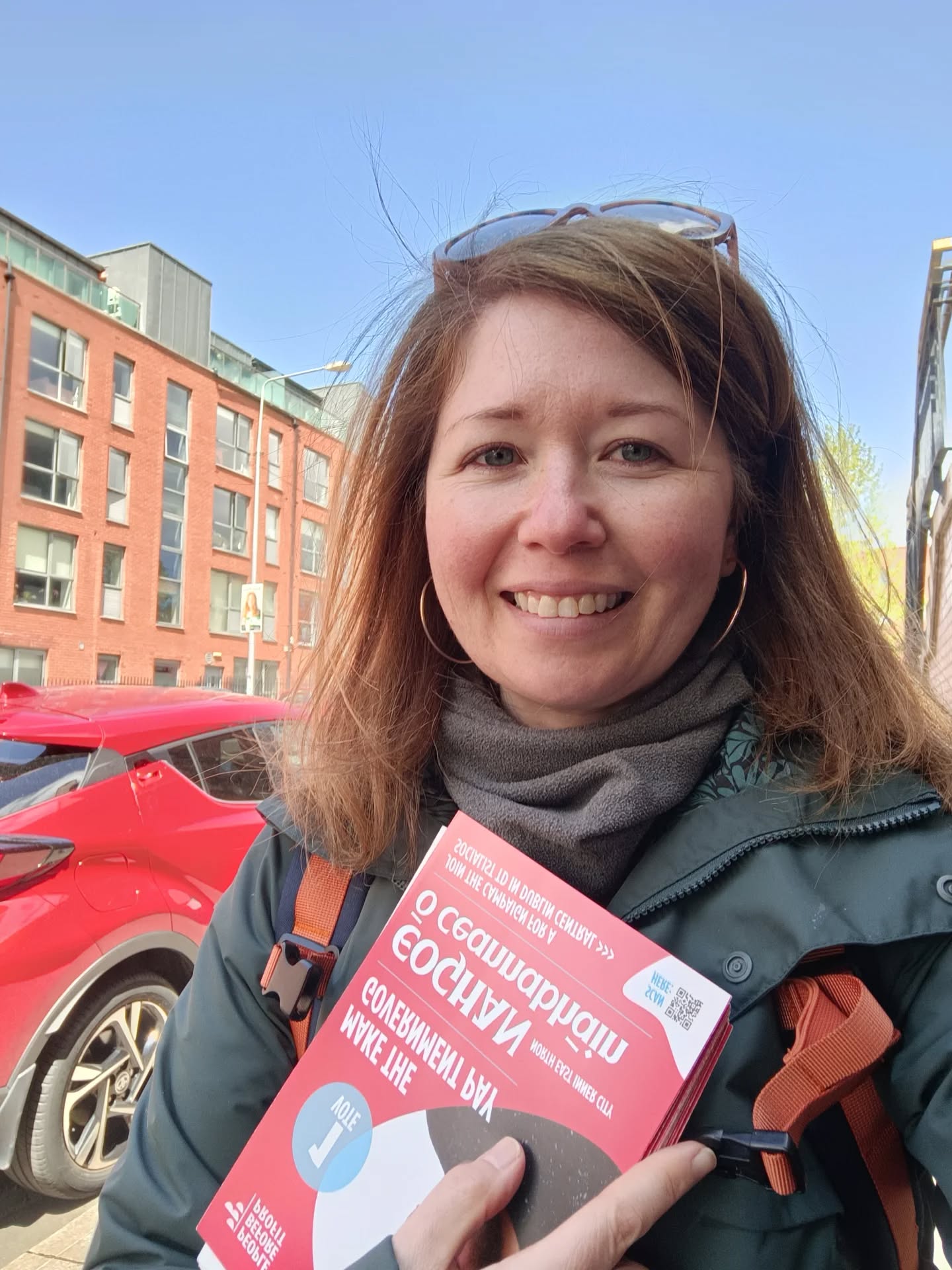 A white women in her 30s or 40s smiles at the camera on the street on a sunny day. 