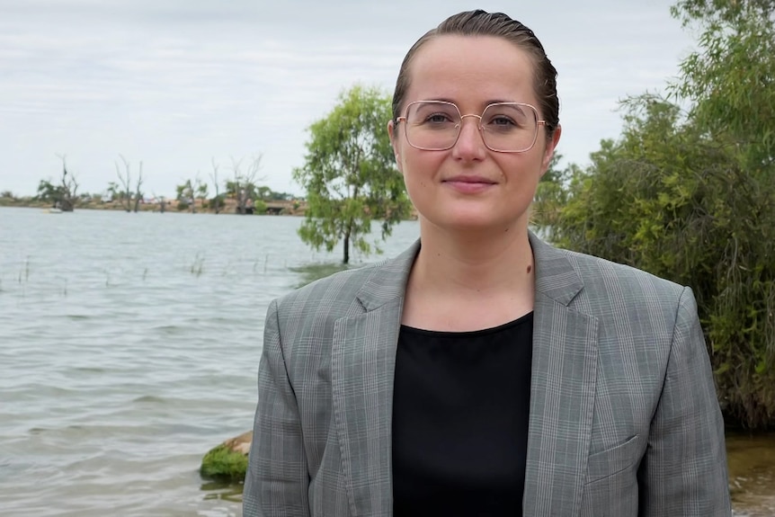 A white woman with brown hair wearing a black shirt and grey coat in front of a body of water and green shrubs.