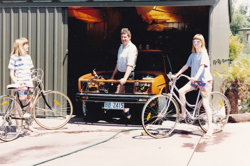 Two teens on bicycles look at their dad, who stands in a yellow cars engine cavity.