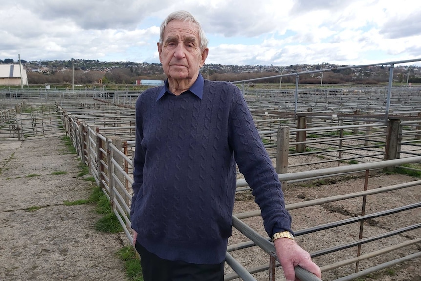 an elderly man stands against a row of steel pens that normally hold sheep