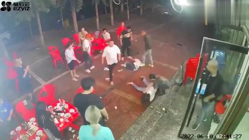 Two women lie on the ground as a group of men surround them outside a restaurant.