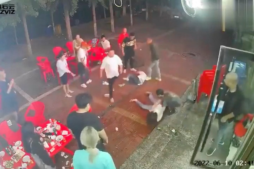 Two women lie on the ground as a group of men surround them outside a restaurant.